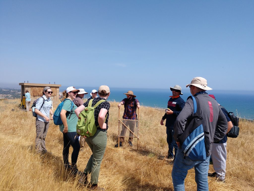 The group listens to a presentation. The sea is visible in the background.
