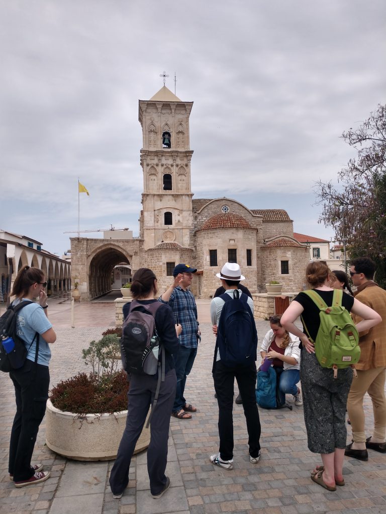 The group stands informally in front of a light-colored stone church with a prominent square bell tower.