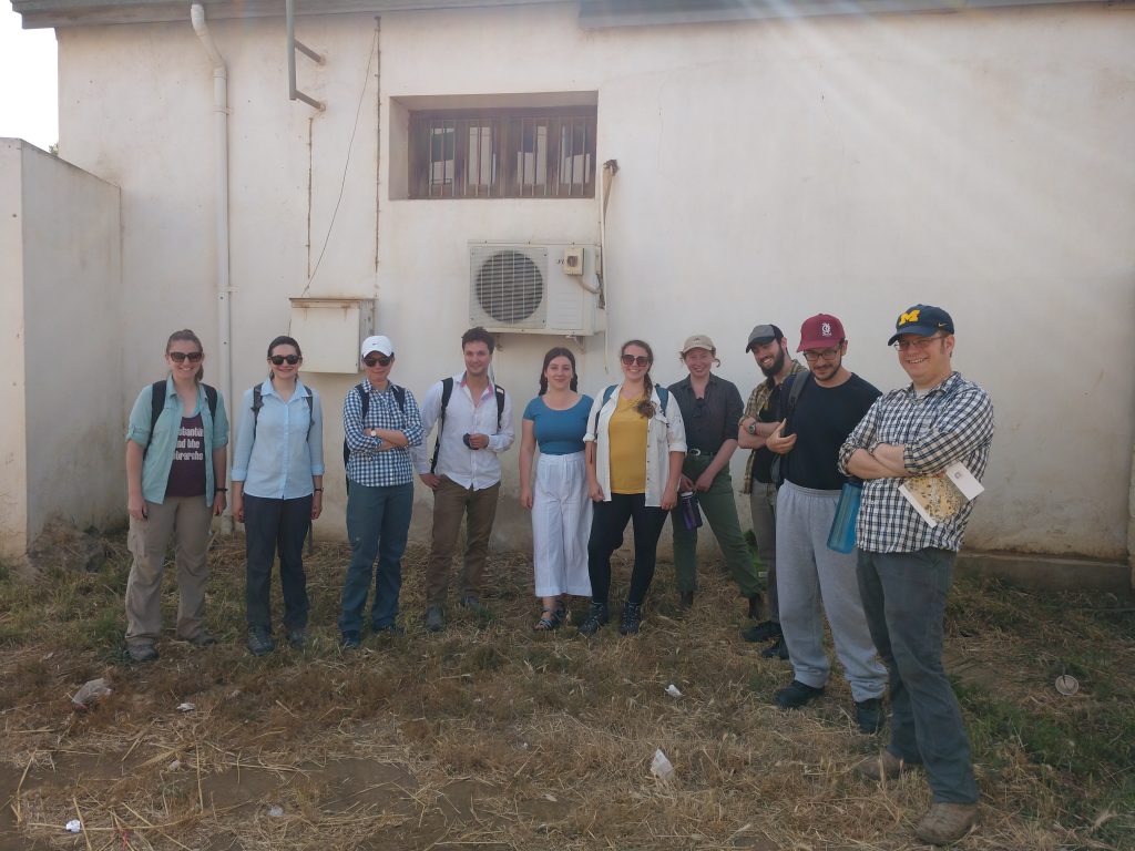 A line of people stand, facing forward, in a weedy lot in front of a dusty building.