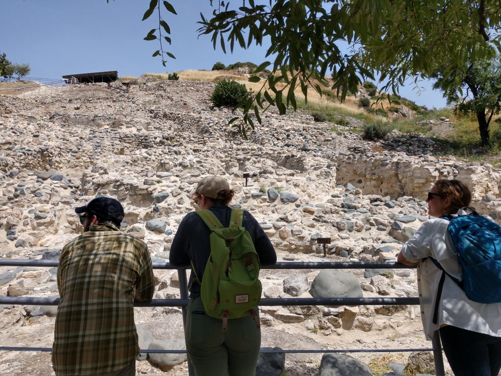 Three people, facing away from the camera, lean against a metal fence looking up at a hillside of large rocks, some of which are arranged in circular shapes.