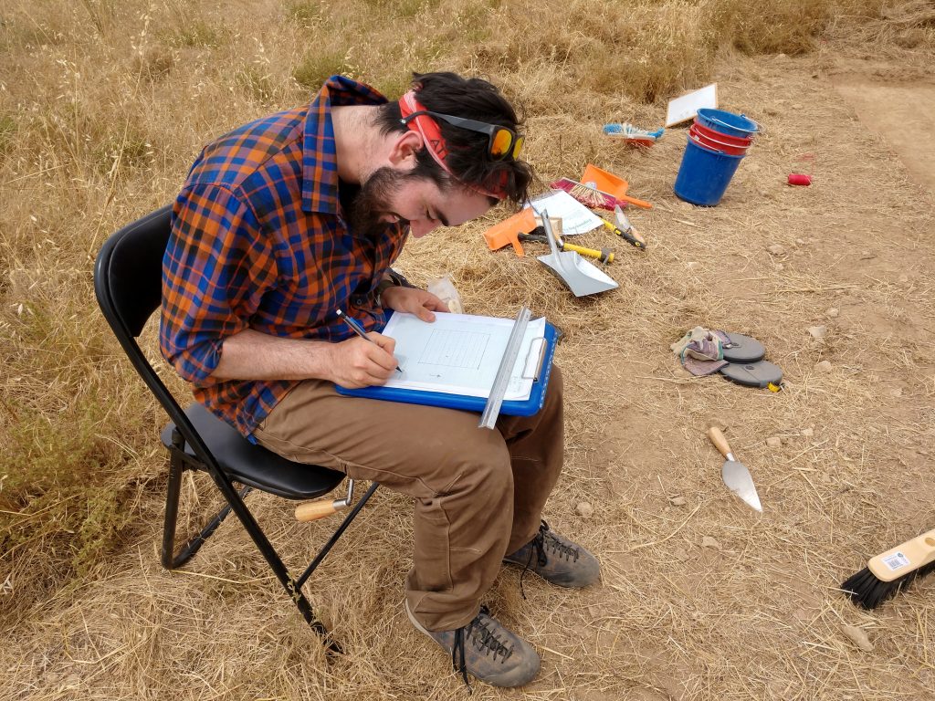 A man sits on a chair in a field among digging tools writing on a clipboard