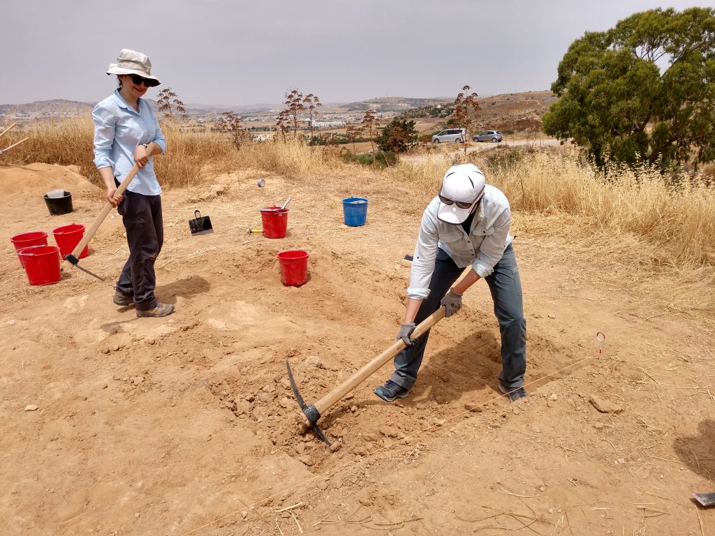 A woman uses a pickax in a dirt field while another woman moves equipment