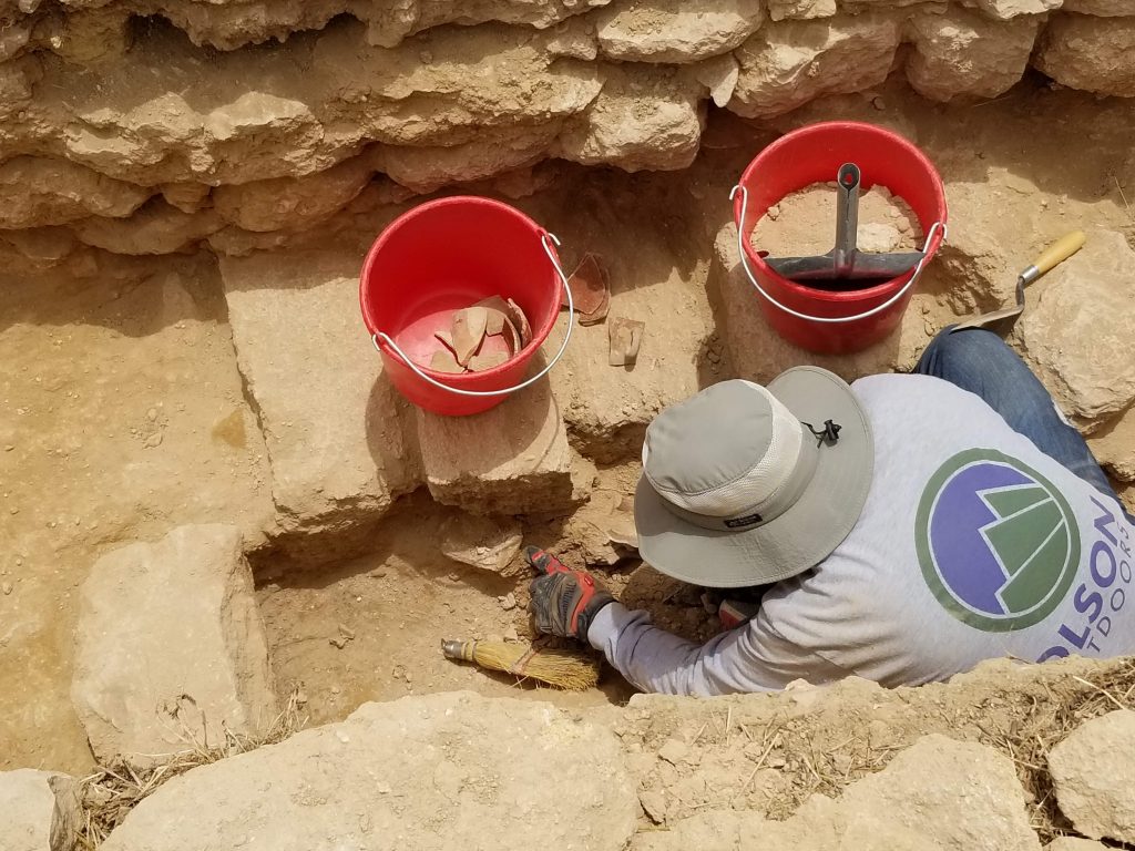 An archaeologist works near an ancient wall filling buckets with dirt and broken pottery