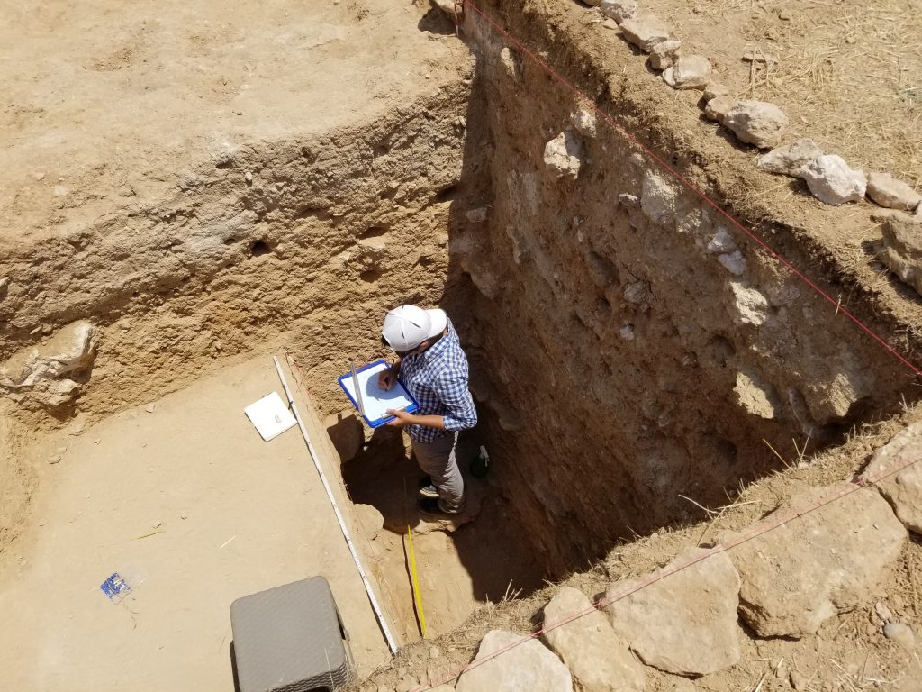 An archaeologist stands in a deep trench drawing