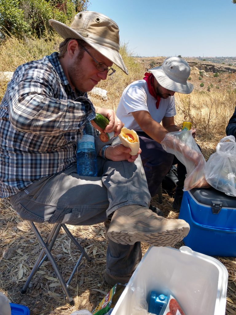 A man eats a sandwich of potato chips, salami, and hot sauce outdoors