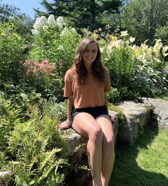 Peri Joy smiling and sitting on a rock wall in a flower garden.