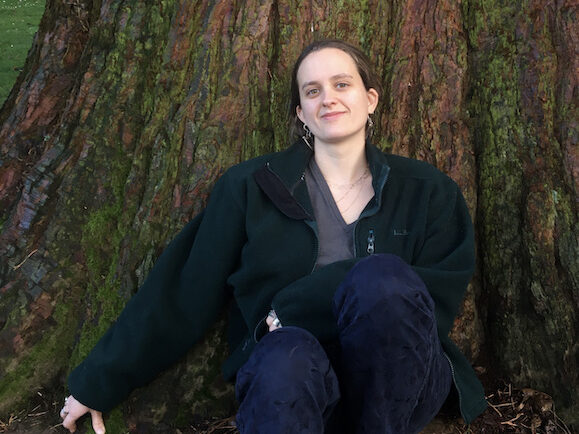 Sofie is sitting up against the trunk of a large redwood tree and looking into the camera with a partial smile.