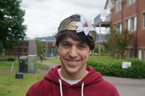 Nate is smiling and wearing plastic laurels with a white flower, and standing on Reed campus, with a dorm building in the background.