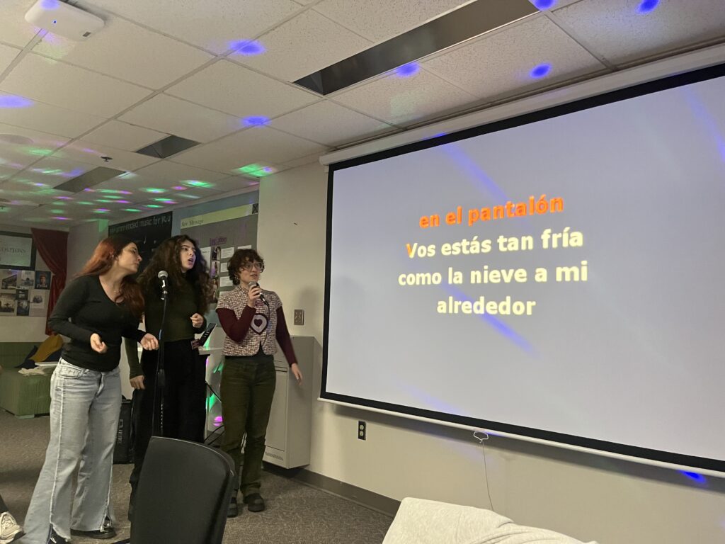 Three students gathered around a microphone in front of a projector screen, singing a song in Spanish.