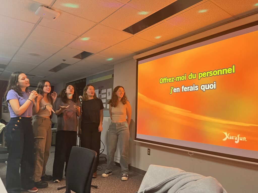Five students gathered around a microphone in front of a projector screen, singing a song in French.