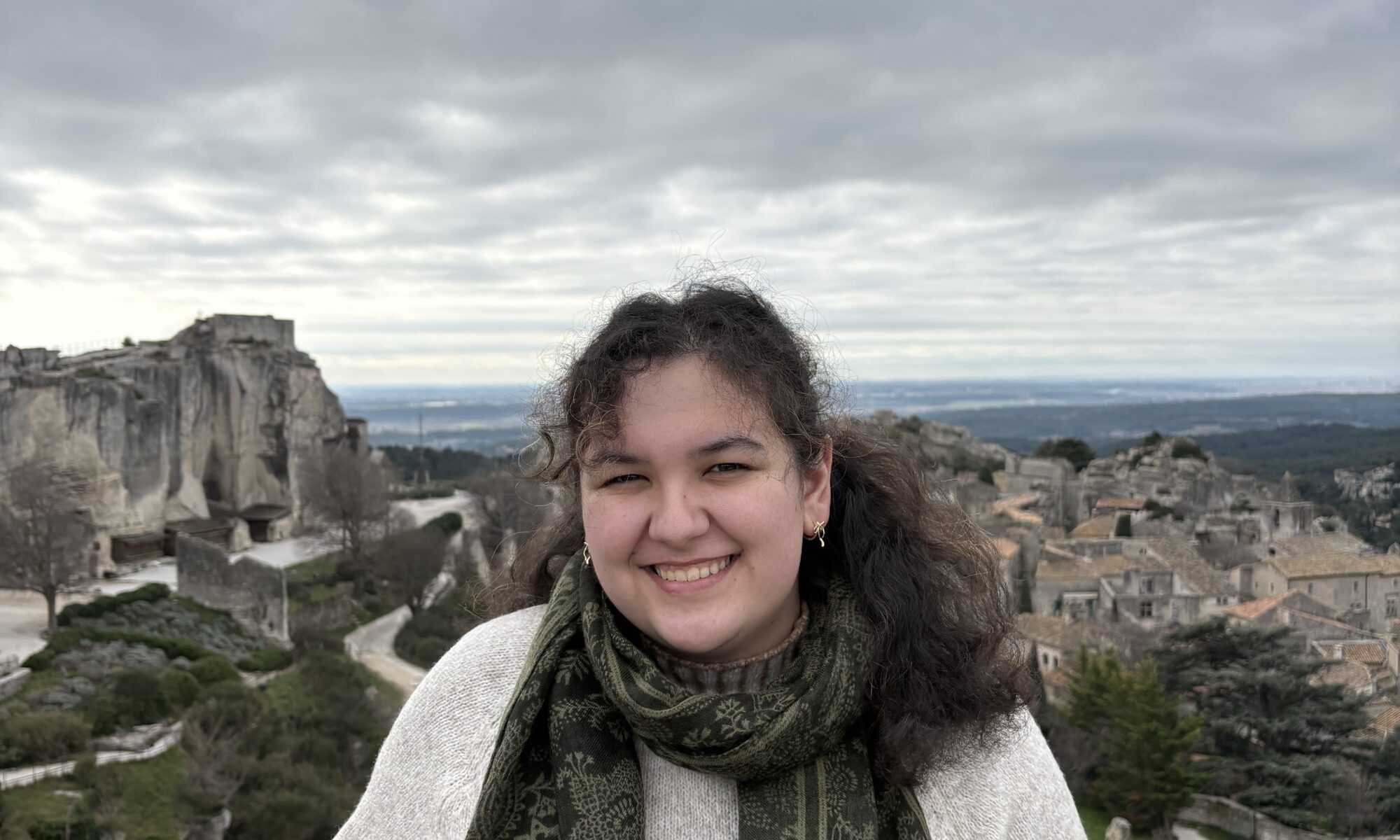 A picture of Miriam smiling. She is in front of a railing, overlooking a city, underneath a high grey sky.
