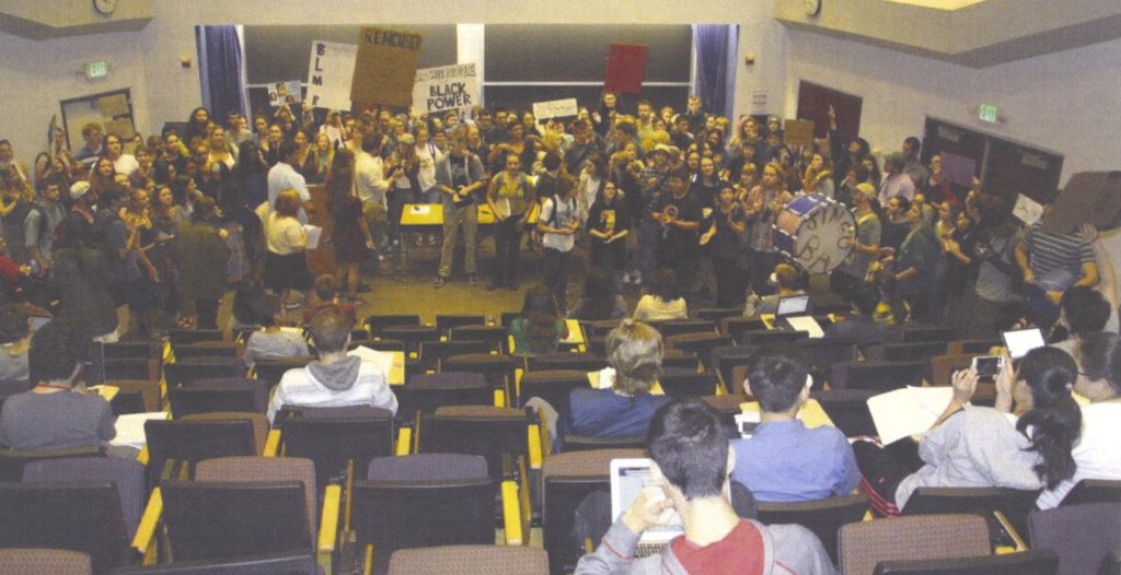 Protestors gathered in a lecture hall holding signs, students watch from the audience. 