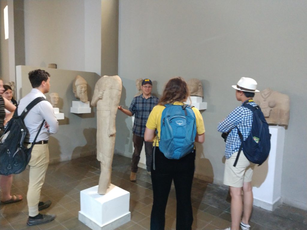 A group of students stands among ancient sculptures of at a museum